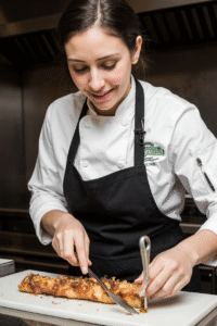 A female chef in a white coat and black apron slicing a baked dish in a professional kitchen.